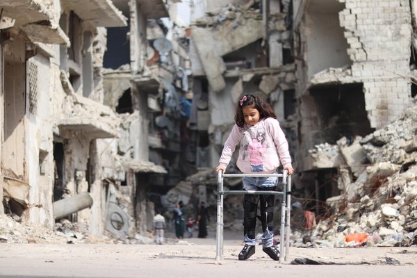 A girl with a disability is standing in front of a shattered building.